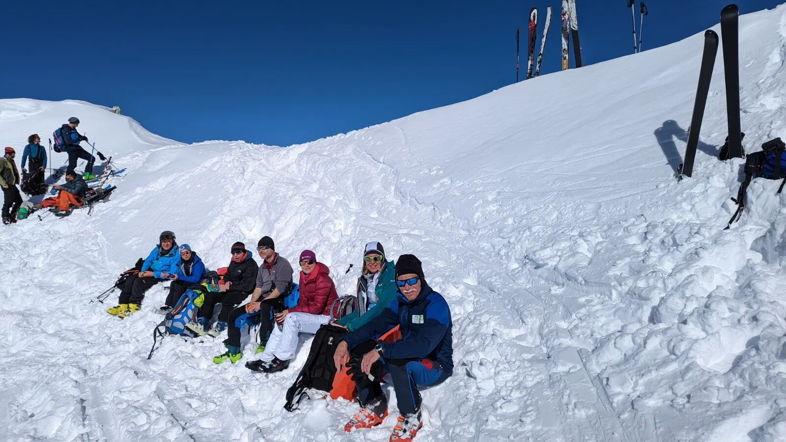 Sieben Personen machen bei Sonnenschein eine Pause im Schnee, Skier stehen neben ihnen im Hang. | © DAV Dortmund