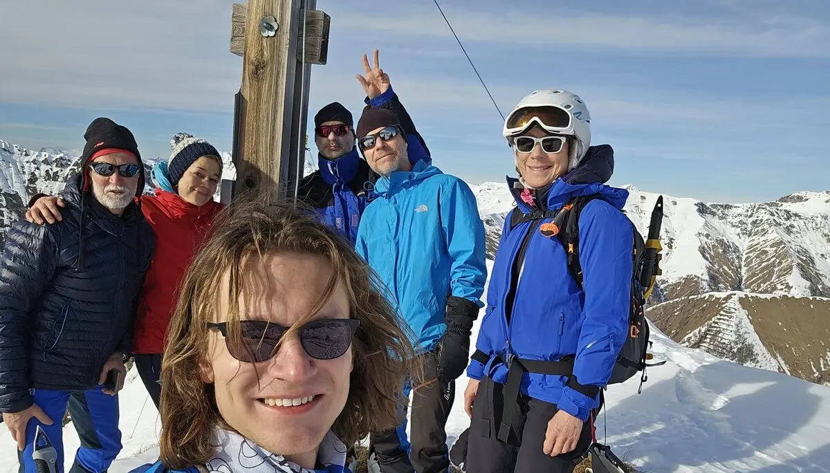 Sechs Skitourengeher*innen machen ein Selfie an einem Gipfelkreuz bei Sonnenschein. | © DAV Dortmund