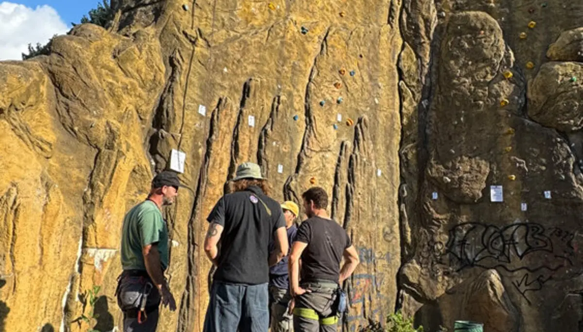 Eine Gruppe von fünf Kletterern steht am Fuß des Kletterturms in Dortmund. Sie besprechen gemeinsam die Routen an der Kletterwand, die mit bunten Griffen und ausgehängten Zetteln markiert ist. | © DAV Dortmund