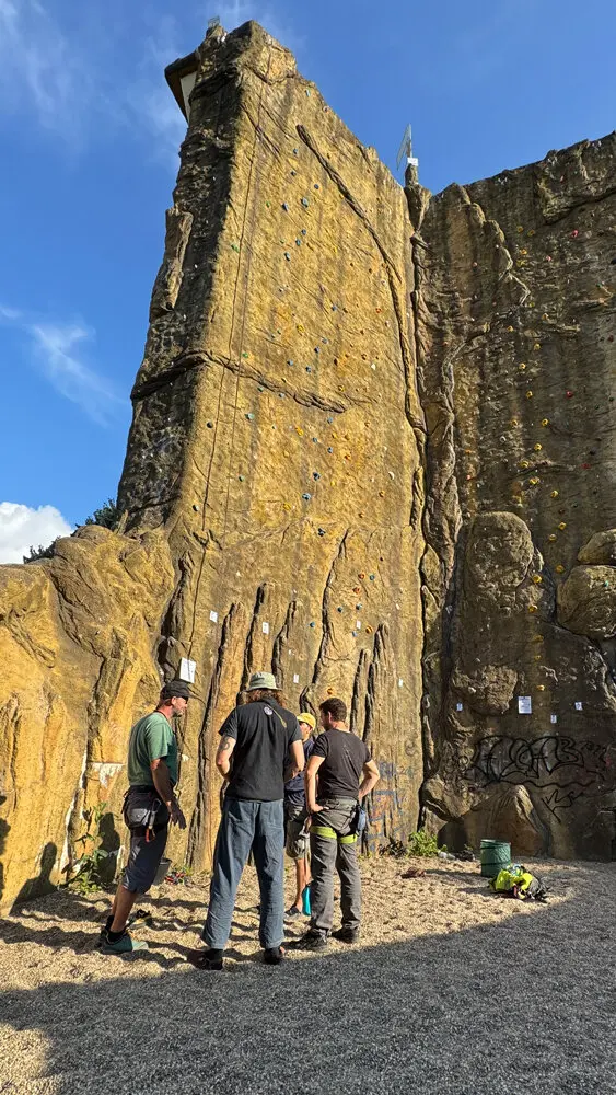 Eine Gruppe von fünf Kletterern steht am Fuß des Kletterturms in Dortmund. Sie besprechen gemeinsam die Routen an der Kletterwand, die mit bunten Griffen und ausgehängten Zetteln markiert ist. | © DAV Dortmund