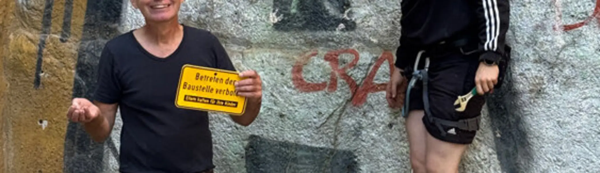 Zwei Männer stehen vor einer bemalten Wand am Kletterturm. Einer hält ein gelbes Schild mit der Aufschrift „Betreten der Baustelle verboten“, der andere steht auf einem Hocker und lacht. | © DAV Dortmund