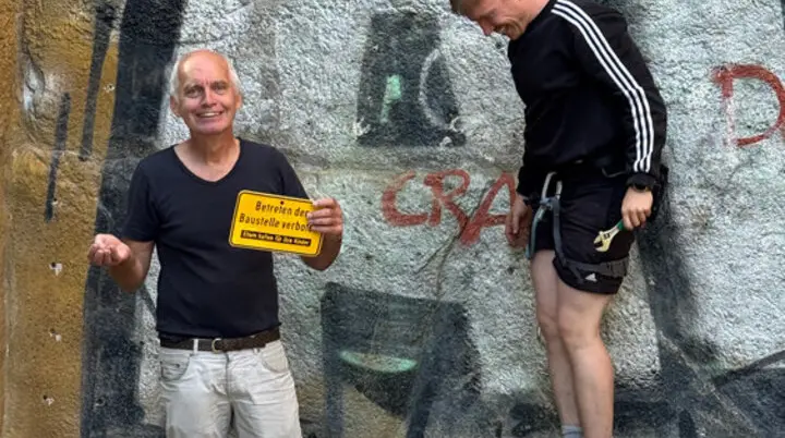 Zwei Männer stehen vor einer bemalten Wand am Kletterturm. Einer hält ein gelbes Schild mit der Aufschrift „Betreten der Baustelle verboten“, der andere steht auf einem Hocker und lacht. | © DAV Dortmund