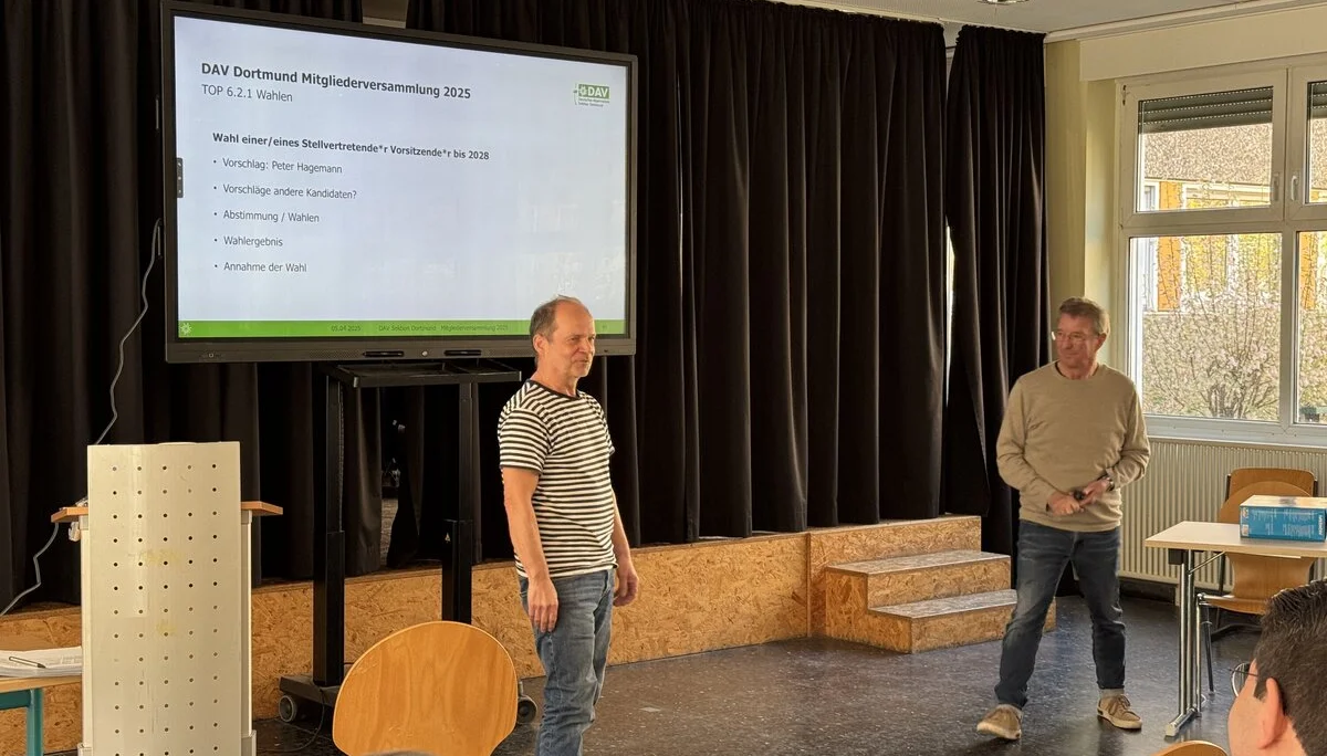 Zwei Männer stehen auf einer kleinen Bühne vor einem großen Bildschirm, auf dem eine Präsentationsfolie mit dem Titel „DAV Dortmund Mitgliederversammlung 2024“ angezeigt wird. Einige Personen sitzen im Publikum. Der Raum ist hell und freundlich, mit Peter Hagemann stellt sich vor.t. | © DAV Dortmund