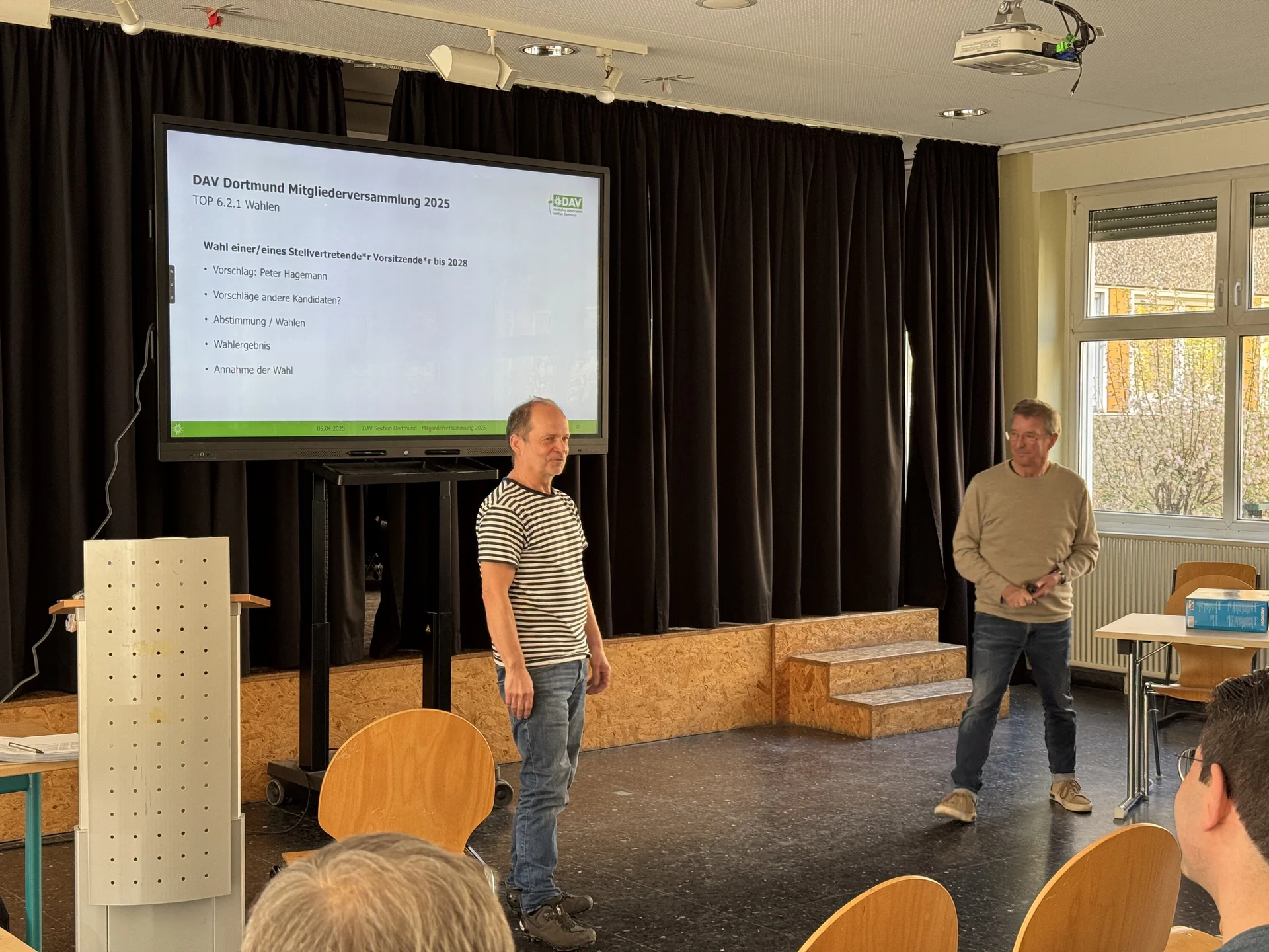 Zwei Männer stehen auf einer kleinen Bühne vor einem großen Bildschirm, auf dem eine Präsentationsfolie mit dem Titel „DAV Dortmund Mitgliederversammlung 2024“ angezeigt wird. Einige Personen sitzen im Publikum. Der Raum ist hell und freundlich, mit Peter Hagemann stellt sich vor.t. | © DAV Dortmund