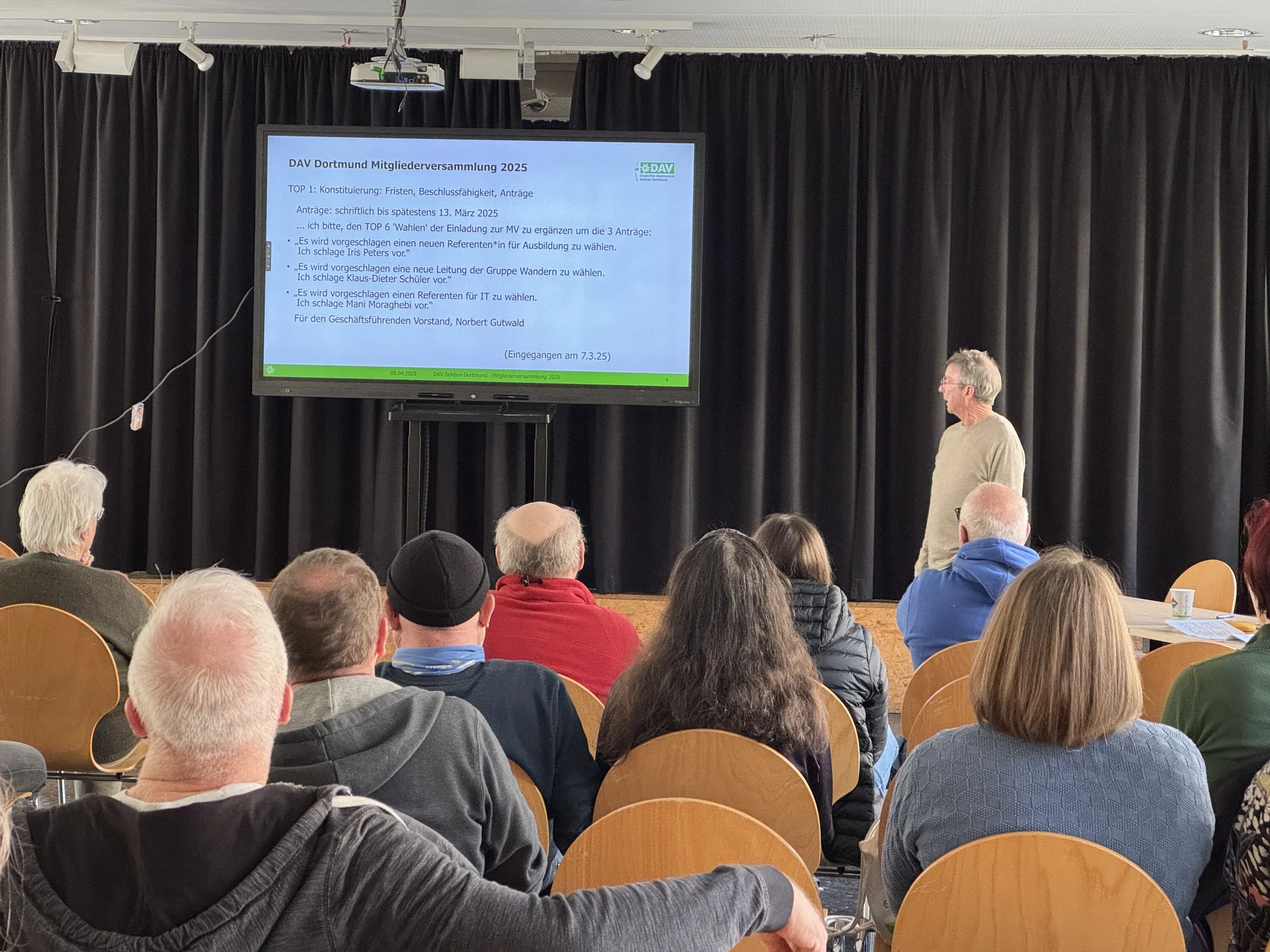 Ein Mann steht vor einer Leinwand mit einer Präsentation über Tagesordnungspunkte bei der DAV Mitgliederversammlung. | © DAV Dortmund