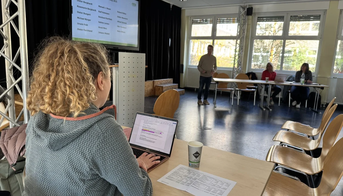 Eine Frau sitzt an einem Tisch mit Laptop und schreibt mit, während im Hintergrund eine Präsentation gezeigt wird. | © DAV Dortmund