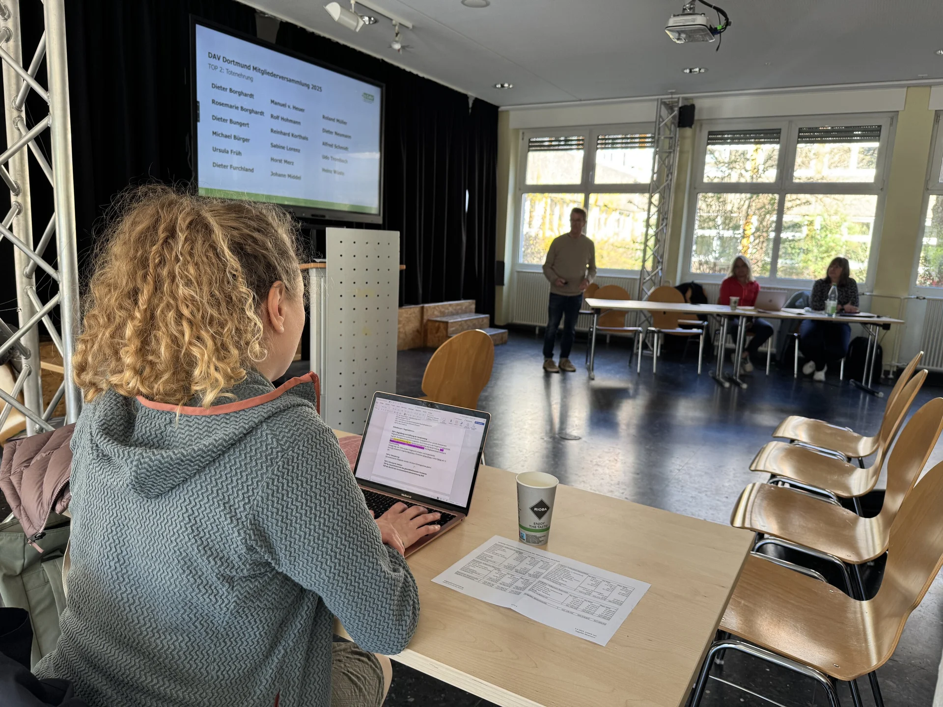 Eine Frau sitzt an einem Tisch mit Laptop und schreibt mit, während im Hintergrund eine Präsentation gezeigt wird. | © DAV Dortmund