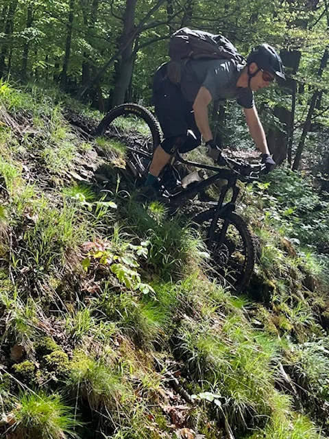 Ein Mountainbiker mit Helm und Rucksack fährt steil bergab durch einen sonnigen Waldabschnitt. | © DAV Dortmund