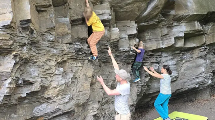 Zwei Kinder klettern nebeneinander an einer Felswand, während drei Erwachsene sie sichern und ein Crashpad am Boden liegt. | © DAV Dortmund