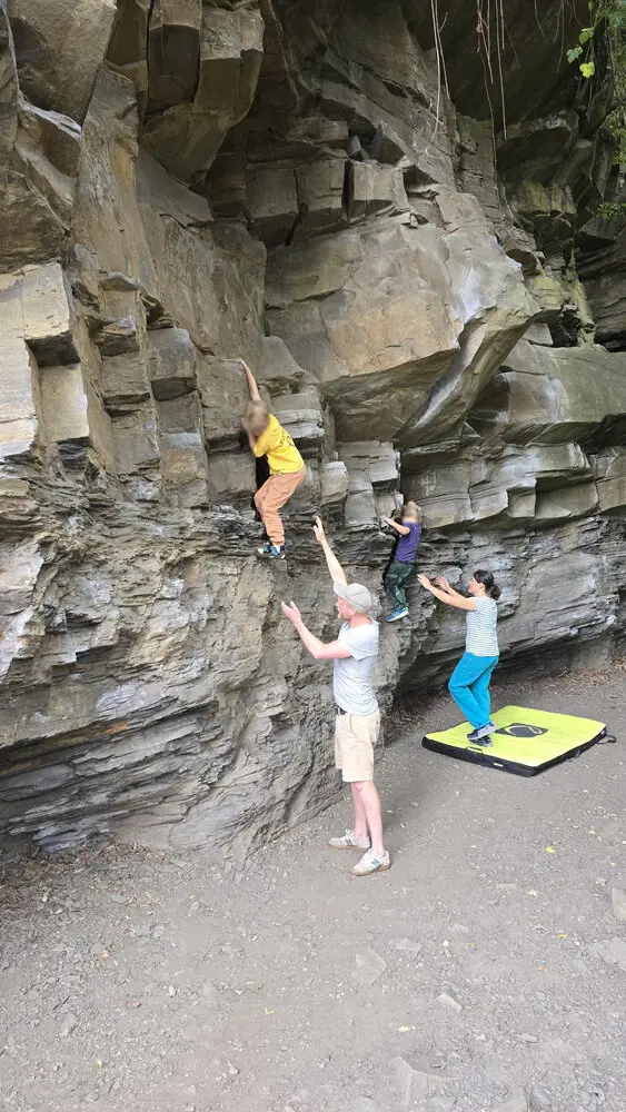 Zwei Kinder klettern nebeneinander an einer Felswand, während drei Erwachsene sie sichern und ein Crashpad am Boden liegt. | © DAV Dortmund
