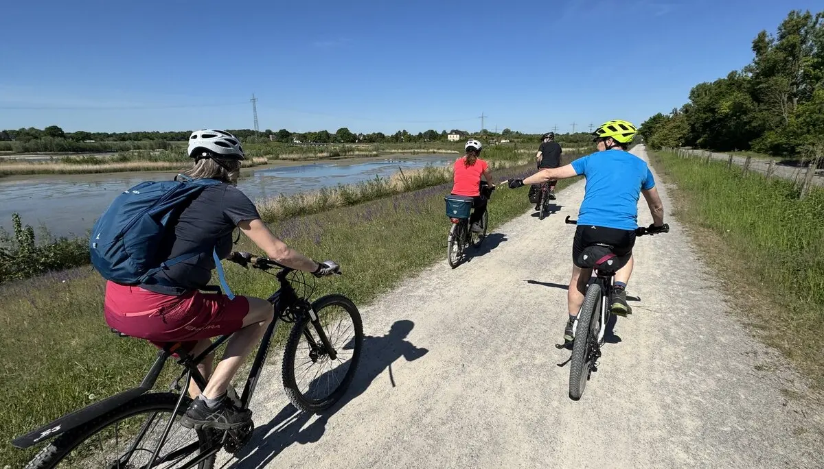 Mehrere Radfahrer*innen fahren bei Sonnenschein auf einem breiten Schotterweg neben einem Gewässer. | © DAV Dortmund