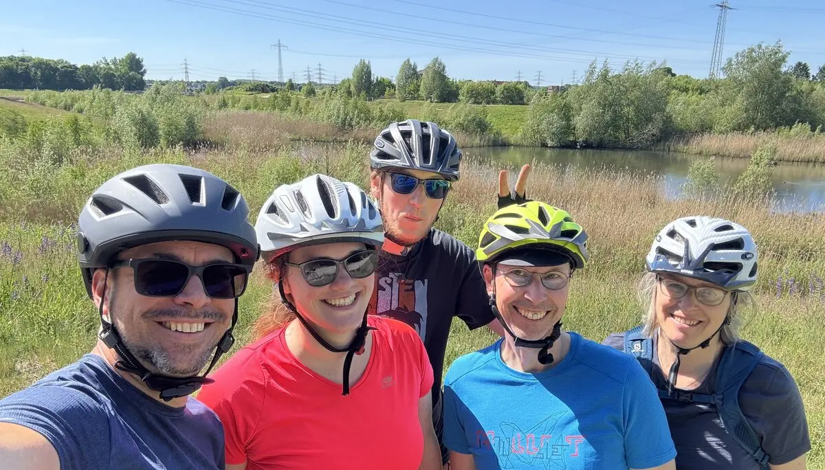 Fünf Radfahrer*innen mit Helm lächeln bei Sonnenschein in die Kamera, im Hintergrund Wiese und Stromleitungen. | © DAV Dortmund