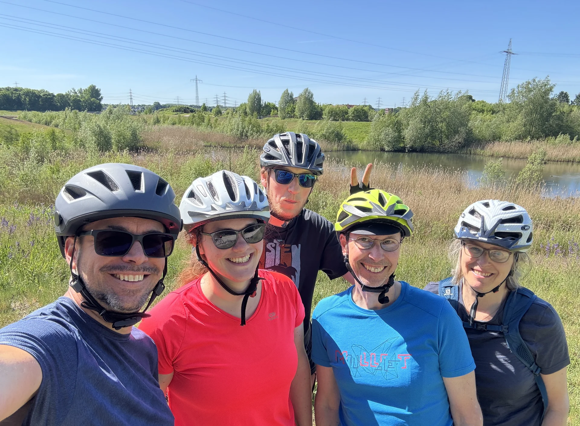 Fünf Radfahrer*innen mit Helm lächeln bei Sonnenschein in die Kamera, im Hintergrund Wiese und Stromleitungen. | © DAV Dortmund