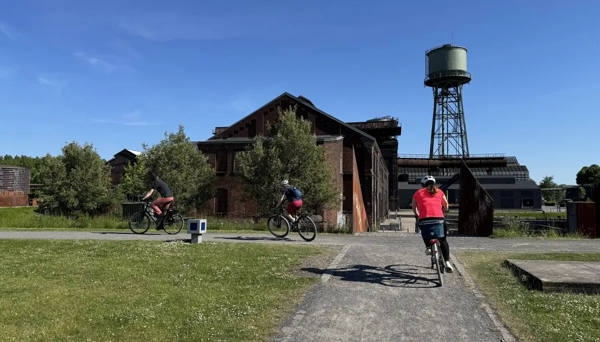 Drei Radfahrer*innen fahren durch ein ehemaliges Industriegelände mit alten Ziegelgebäuden und Wasserturm. | © DAV Dortmund
