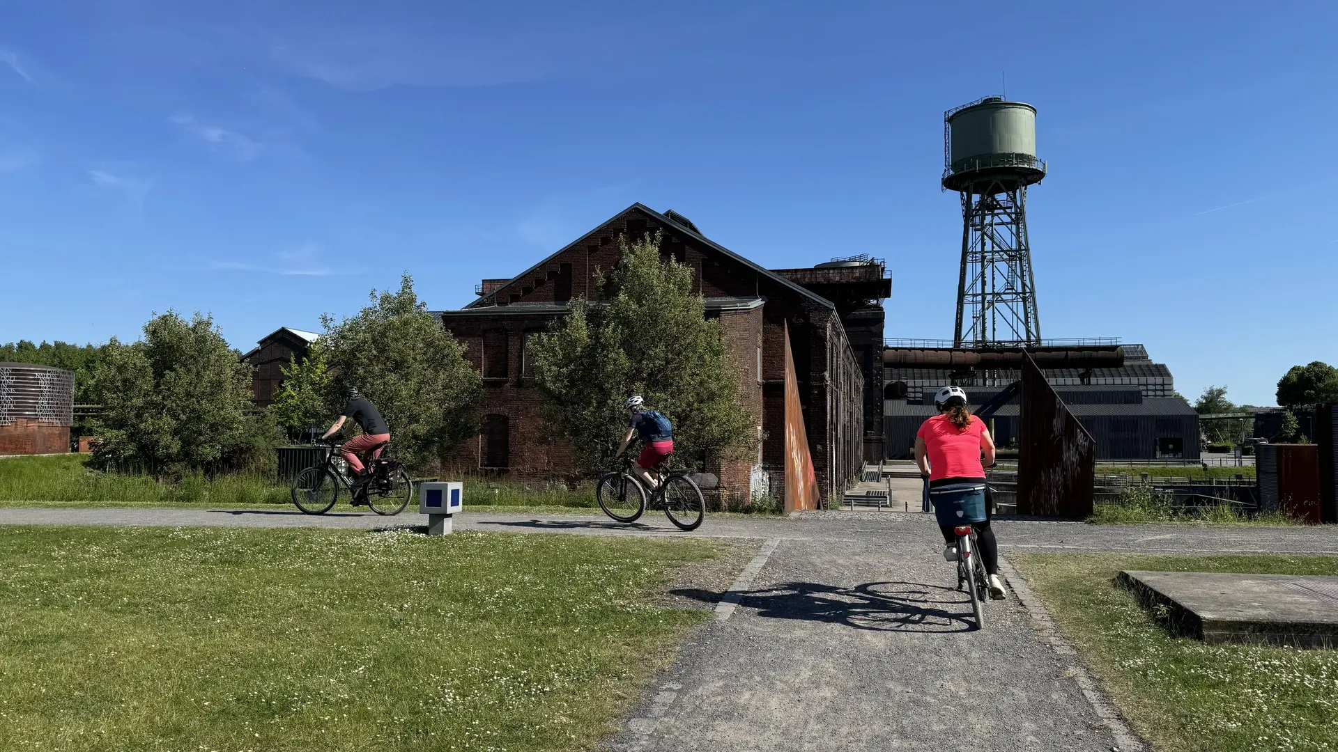 Drei Radfahrer*innen fahren durch ein ehemaliges Industriegelände mit alten Ziegelgebäuden und Wasserturm. | © DAV Dortmund