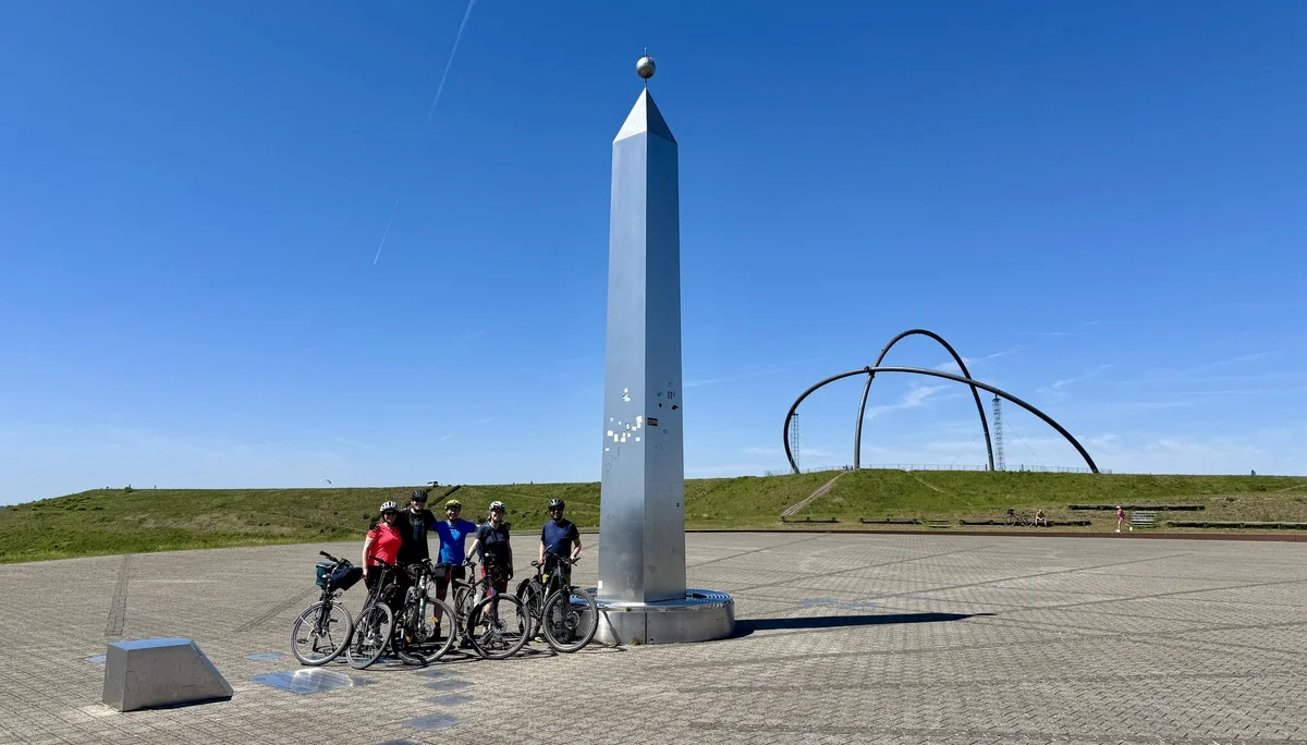 Fünf Radfahrer*innen stehen mit ihren Fahrrädern vor einem großen silbernen Obelisken und Kunstobjekt. | © DAV Dortmund