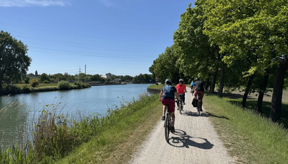 Gruppe radelt auf einem Schotterweg direkt am Wasser unter grünem Blätterdach. | © DAV Dortmund