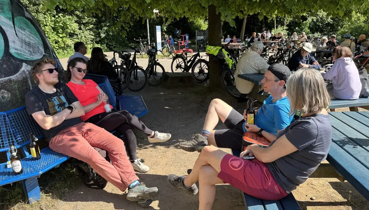 Fünf Personen entspannen mit Getränken auf Bänken unter Bäumen, im Hintergrund stehen viele Fahrräder. | © DAV Dortmund