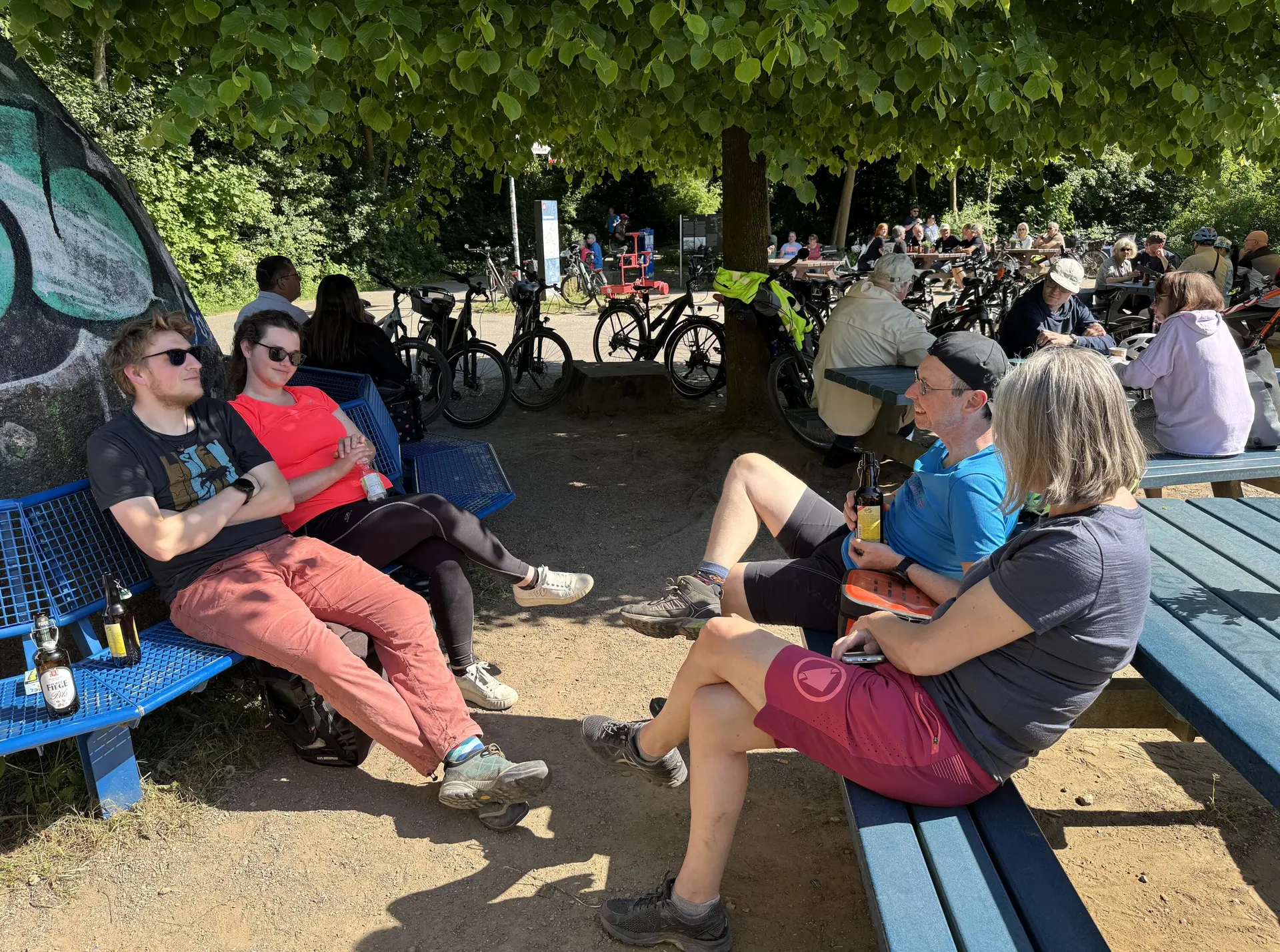 Fünf Personen entspannen mit Getränken auf Bänken unter Bäumen, im Hintergrund stehen viele Fahrräder. | © DAV Dortmund