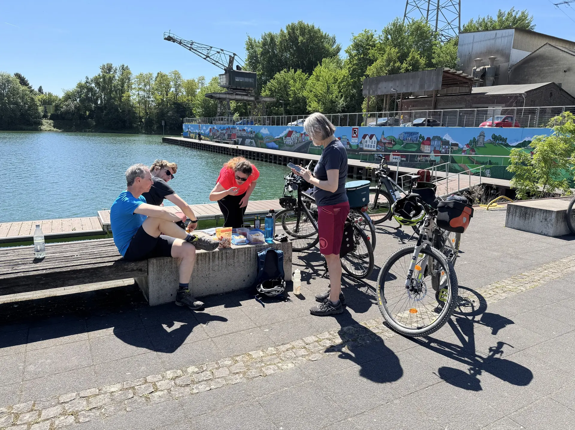 Vier Personen mit Fahrrädern sitzen und stehen an einem Ufer, essen und trinken bei Sonnenschein. | © DAV Dortmund