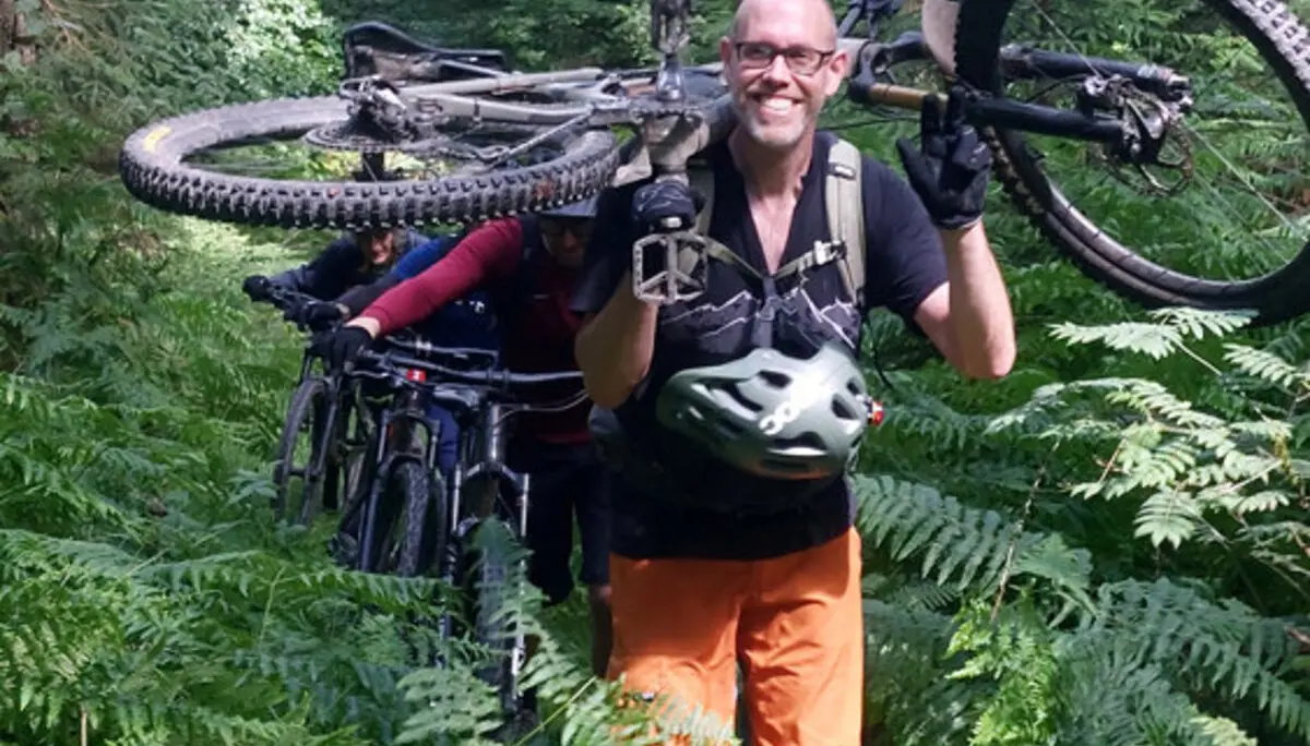 Ein lächelnder Mountainbiker trägt sein Fahrrad durch dichten Farn im Wald. Weitere Biker folgen ihm mit ihren Rädern. | © DAV Dortmund