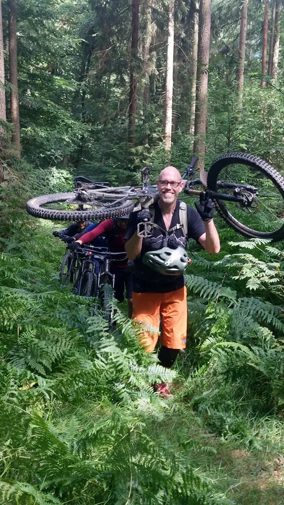 Ein lächelnder Mountainbiker trägt sein Fahrrad durch dichten Farn im Wald. Weitere Biker folgen ihm mit ihren Rädern. | © DAV Dortmund