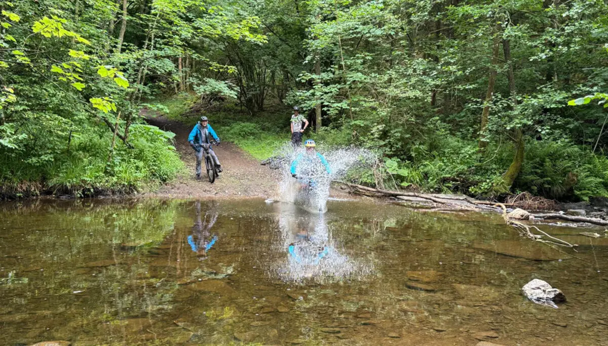 Eine Mountainbikerin fährt mit Schwung durch eine flache Wasserstelle im Wald, während zwei andere zuschauen. Große Wasserspritzer sind zu sehen. | © DAV Dortmund