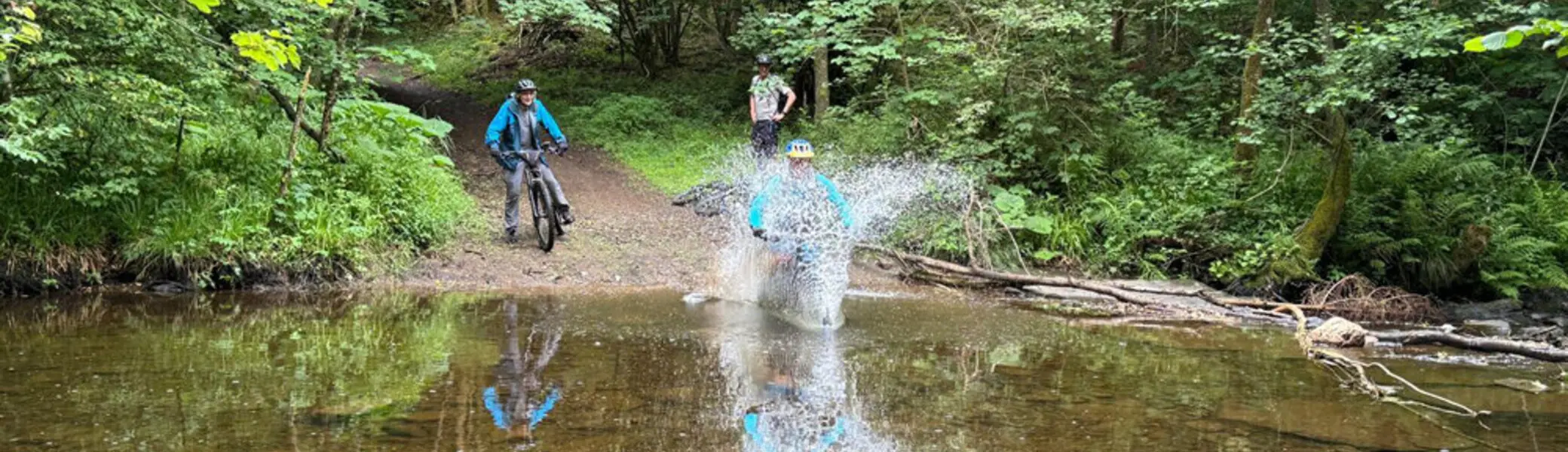 Eine Mountainbikerin fährt mit Schwung durch eine flache Wasserstelle im Wald, während zwei andere zuschauen. Große Wasserspritzer sind zu sehen. | © DAV Dortmund