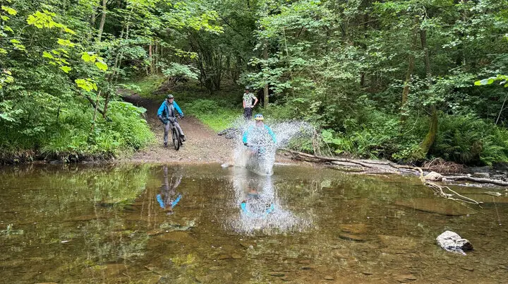 Eine Mountainbikerin fährt mit Schwung durch eine flache Wasserstelle im Wald, während zwei andere zuschauen. Große Wasserspritzer sind zu sehen. | © DAV Dortmund