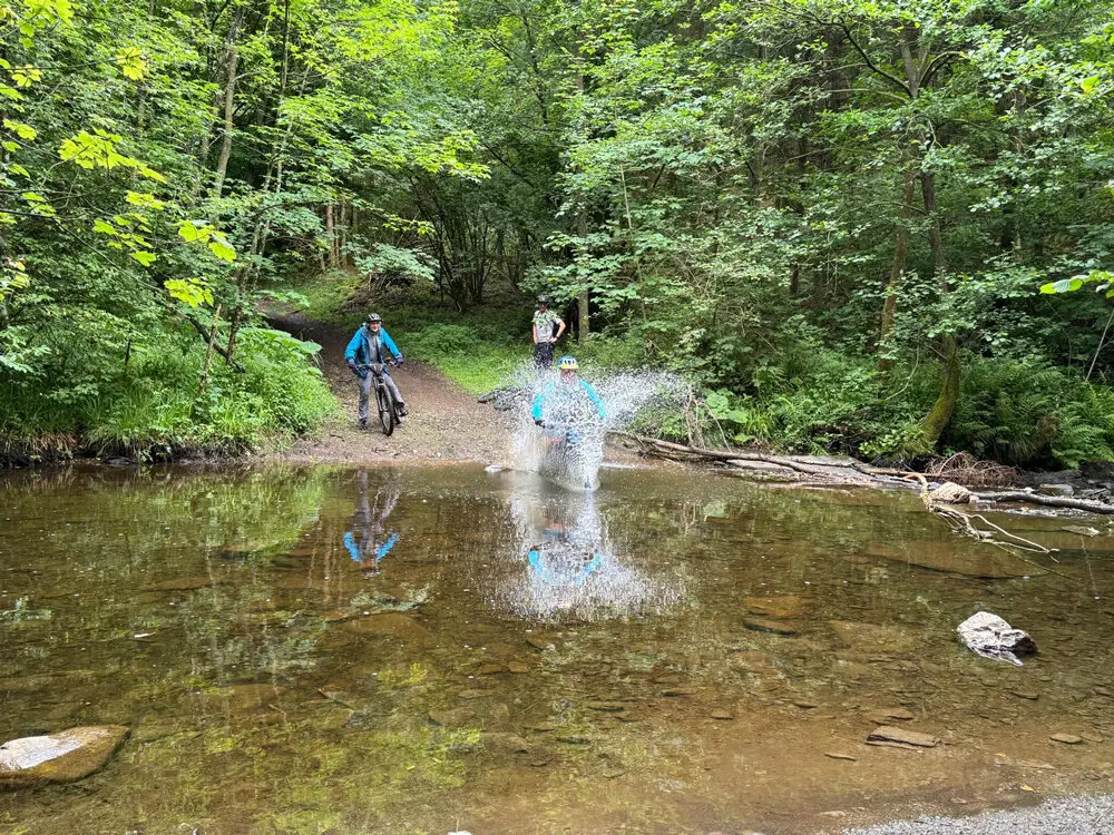 Eine Mountainbikerin fährt mit Schwung durch eine flache Wasserstelle im Wald, während zwei andere zuschauen. Große Wasserspritzer sind zu sehen. | © DAV Dortmund