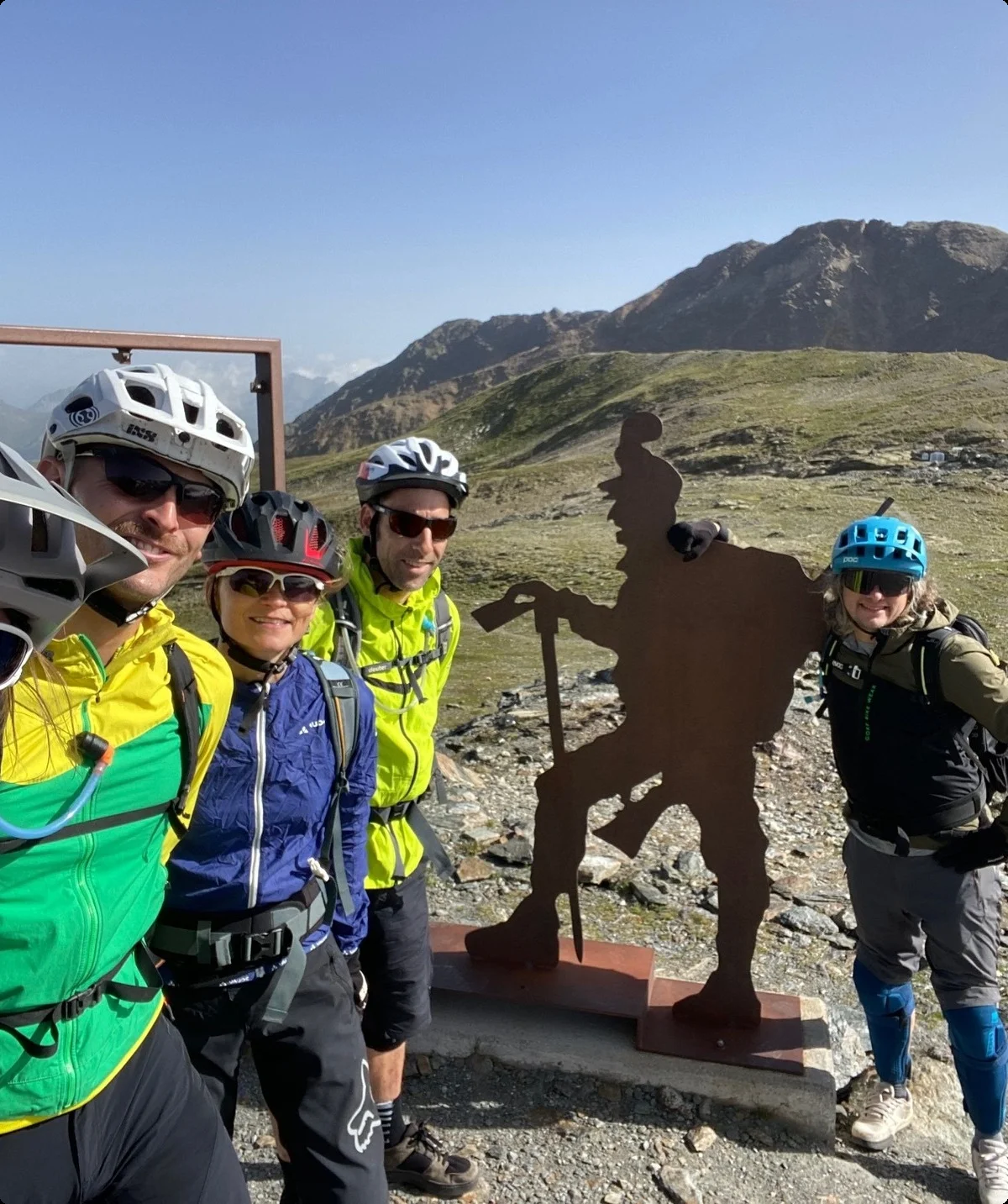 Fünf Mountainbiker posieren an einer Statue mit Bergsteigerfigur vor alpinem Panorama. | © DAV Dortmund