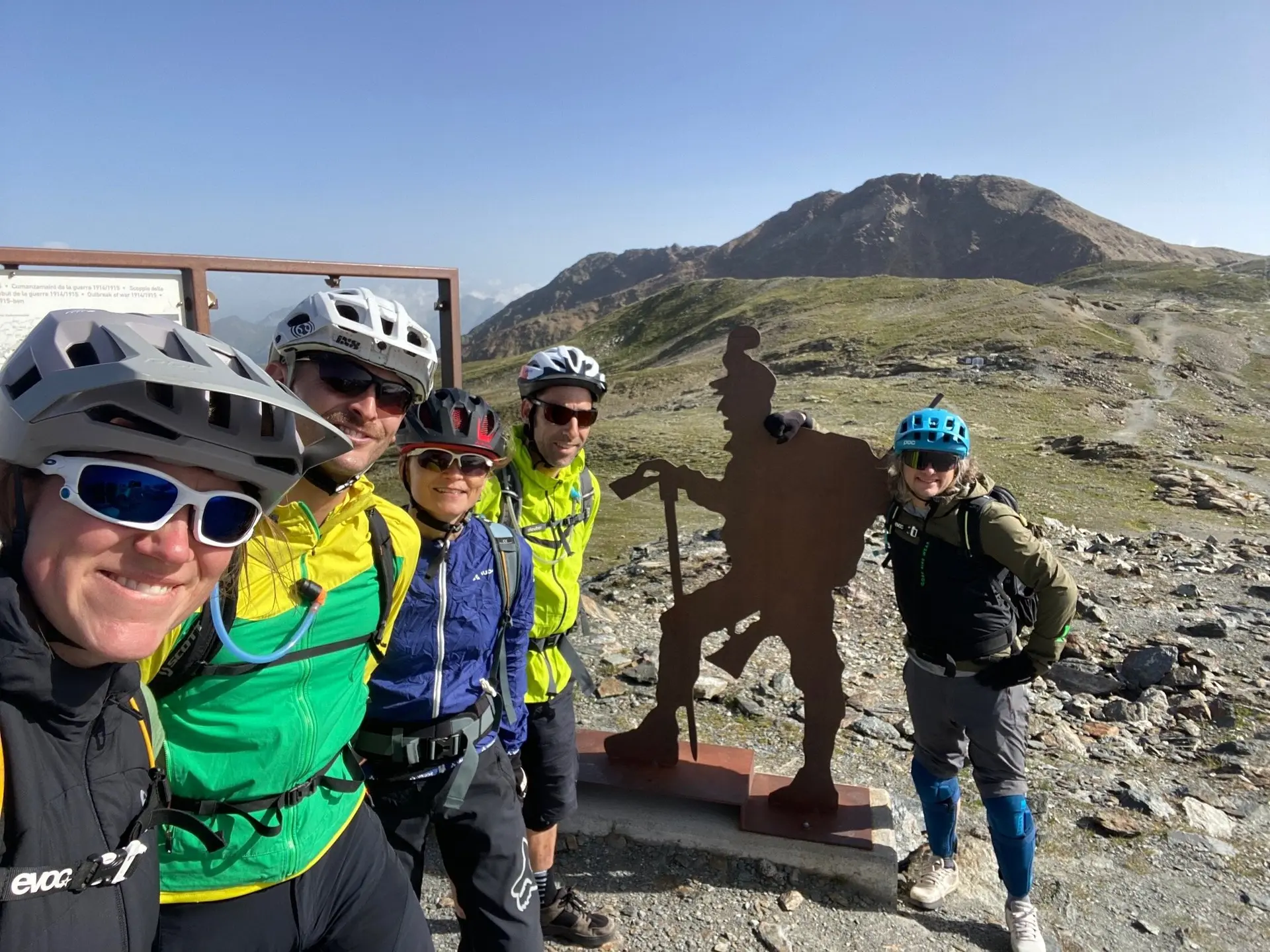 Fünf Mountainbiker posieren an einer Statue mit Bergsteigerfigur vor alpinem Panorama. | © DAV Dortmund