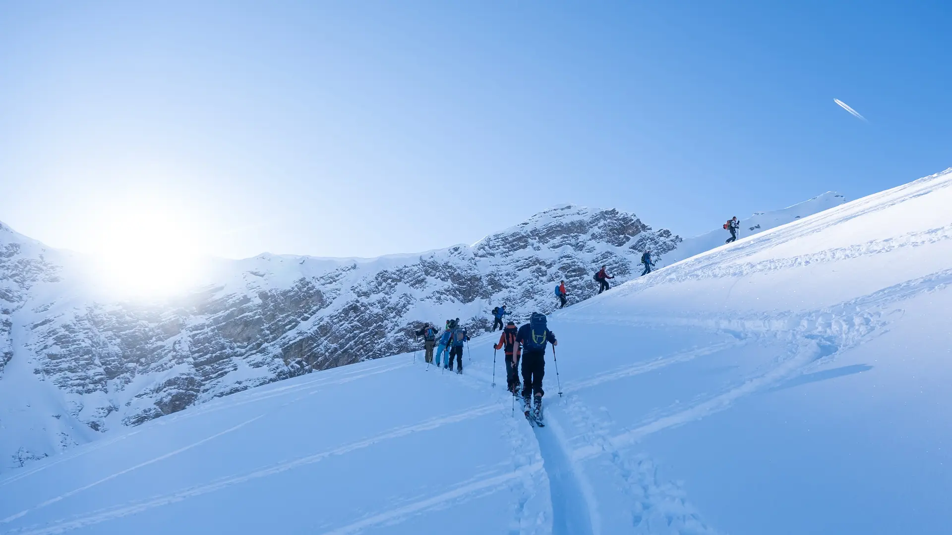 Gruppe von Skitourengeher*innen steigt bei Sonnenaufgang über unberührten Schnee zum Berg auf. | © Fabian Monzigemba