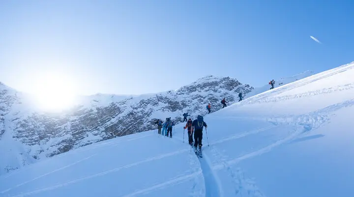 Gruppe von Skitourengeher*innen steigt bei Sonnenaufgang über unberührten Schnee zum Berg auf. | © Fabian Monzigemba
