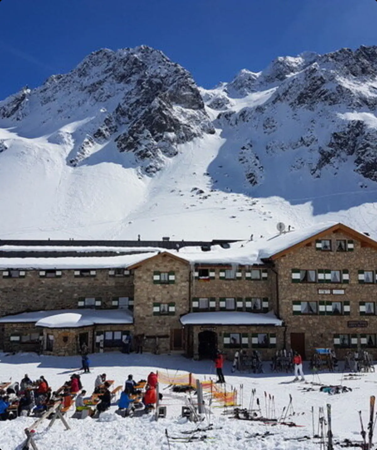 Eine große Skihütte liegt am Hang, umgeben von Schnee. Viele Menschen sitzen auf der sonnigen Terrasse. | © DAV Dortmund