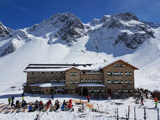 Eine große Skihütte liegt am Hang, umgeben von Schnee. Viele Menschen sitzen auf der sonnigen Terrasse. | © DAV Dortmund