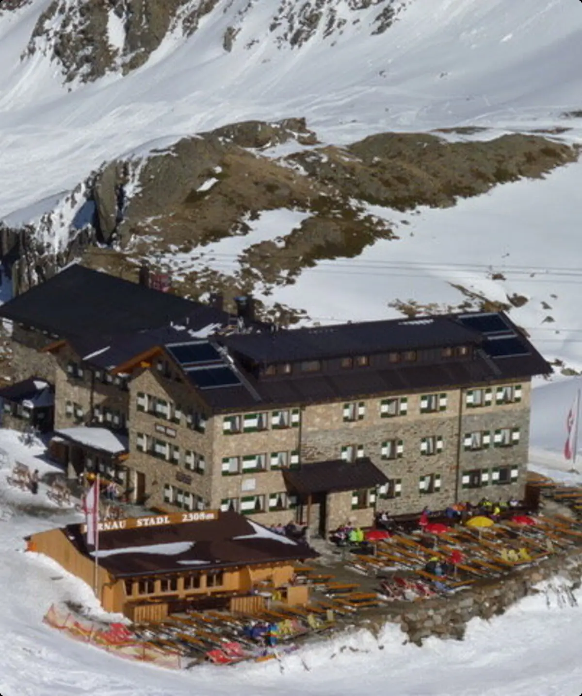 Große Skihütte in verschneiter Berglandschaft, vor der Terrasse stehen viele bunte Liegestühle, Gondeln fahren im Hintergrund. | © DAV Dortmund