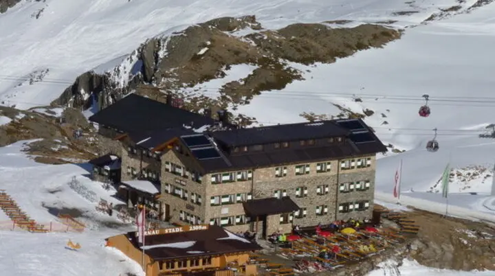 Große Skihütte in verschneiter Berglandschaft, vor der Terrasse stehen viele bunte Liegestühle, Gondeln fahren im Hintergrund. | © DAV Dortmund