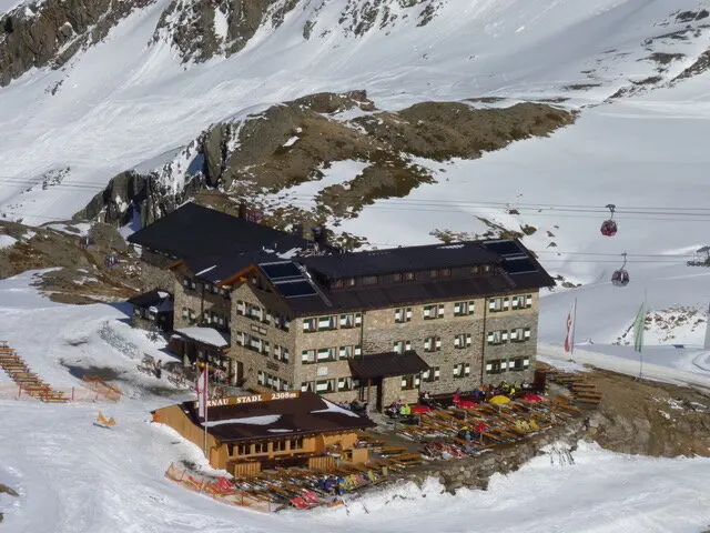 Große Skihütte in verschneiter Berglandschaft, vor der Terrasse stehen viele bunte Liegestühle, Gondeln fahren im Hintergrund. | © DAV Dortmund