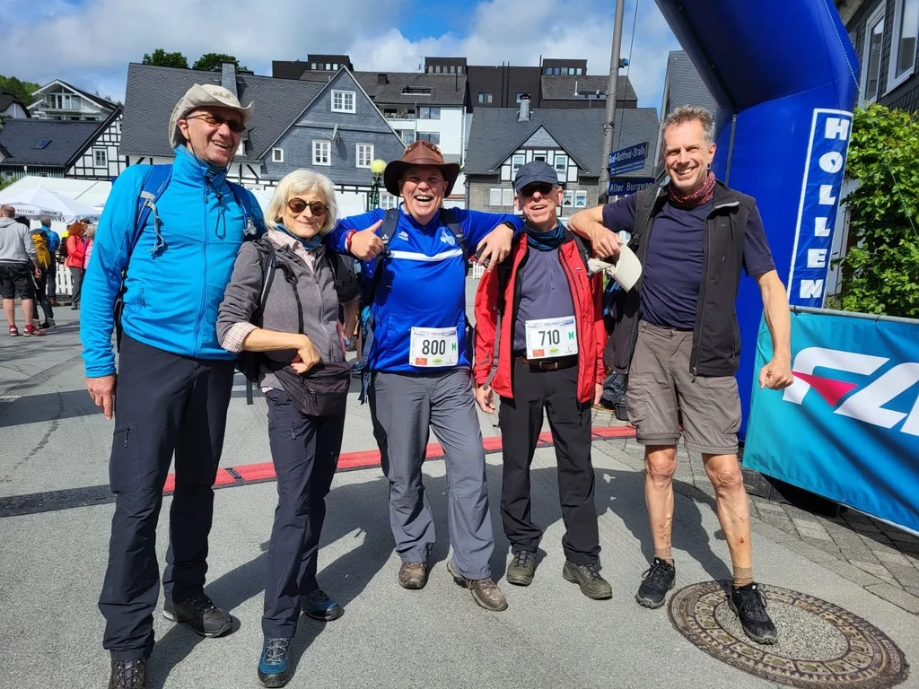 Fünf Personen stehen nach einem Wanderlauf unter dem Zieleinlaufbogen, einige tragen Startnummern und Wanderkleidung. | © DAV Dortmund