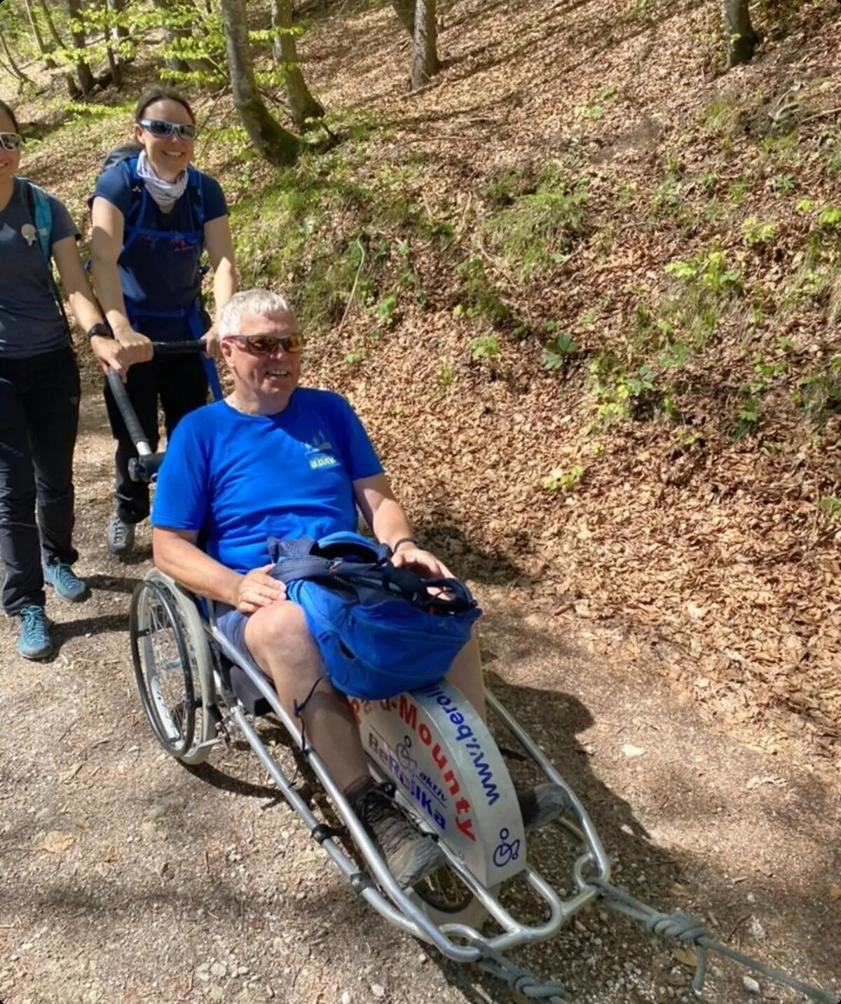  Eine Person sitzt in einem Outdoor-Rollstuhl auf einem Waldweg, zwei Begleiter*innen schieben und helfen. | © DAV Dortmund