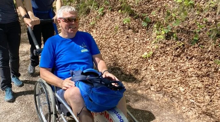  Eine Person sitzt in einem Outdoor-Rollstuhl auf einem Waldweg, zwei Begleiter*innen schieben und helfen. | © DAV Dortmund