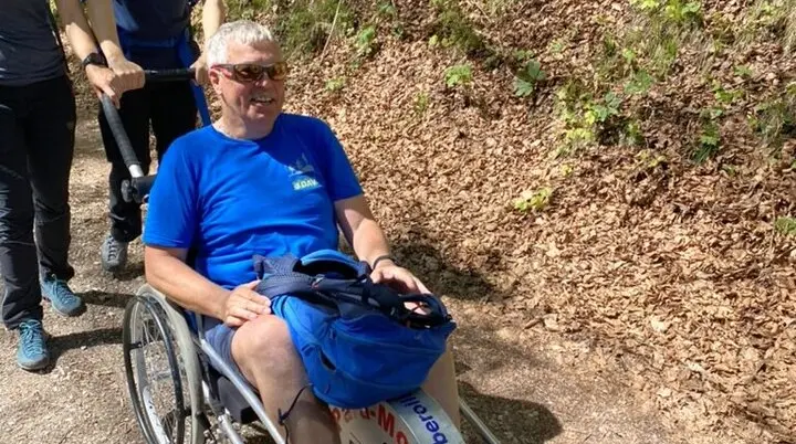  Eine Person sitzt in einem Outdoor-Rollstuhl auf einem Waldweg, zwei Begleiter*innen schieben und helfen. | © DAV Dortmund