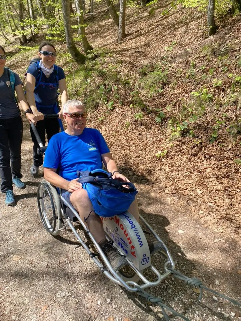  Eine Person sitzt in einem Outdoor-Rollstuhl auf einem Waldweg, zwei Begleiter*innen schieben und helfen. | © DAV Dortmund