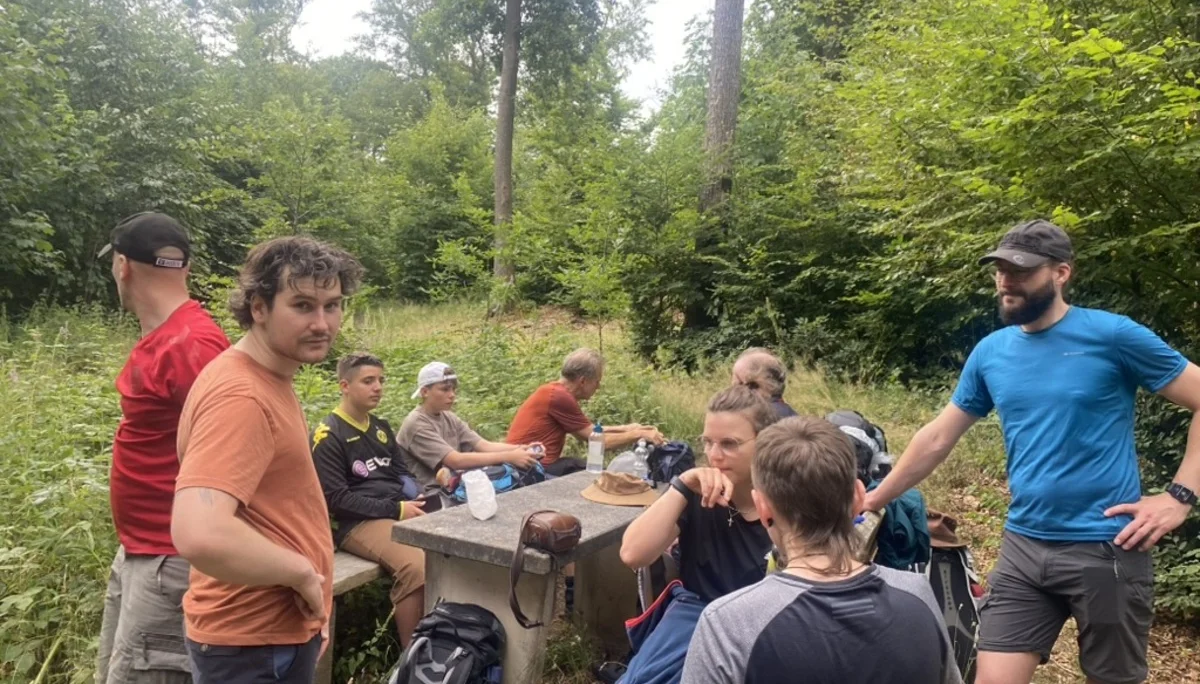 Eine Gruppe von Jugendlichen und Erwachsenen macht Rast an einem Tisch mitten im Wald. Einige stehen, andere sitzen oder lehnen an Rucksäcken. Alle wirken entspannt. | © DAV Dortmund