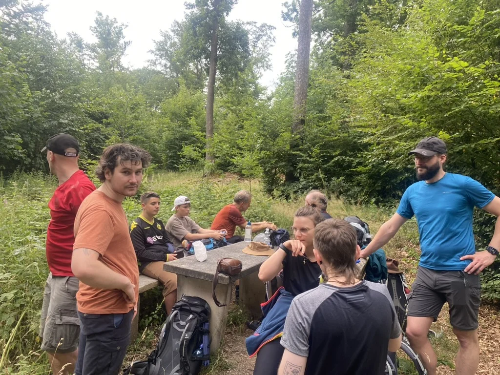 Eine Gruppe von Jugendlichen und Erwachsenen macht Rast an einem Tisch mitten im Wald. Einige stehen, andere sitzen oder lehnen an Rucksäcken. Alle wirken entspannt. | © DAV Dortmund