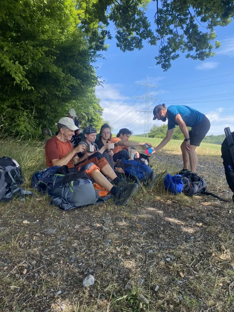 Eine Gruppe von Wandernden sitzt am Waldrand unter einem Baum im Schatten. Sie trinken aus Bechern und entspannen sich auf ihren Rucksäcken. | © DAV Dortmund