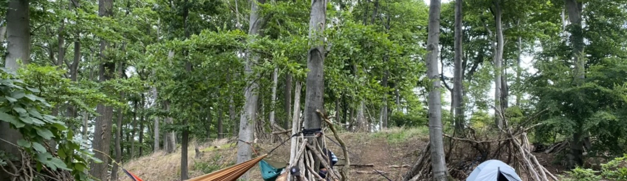 Zwei Hängematten und ein Zelt sind zwischen Bäumen im Wald aufgebaut. Rundherum stehen improvisierte Unterstände aus Ästen und mehrere Rucksäcke. | © DAV Dortmund