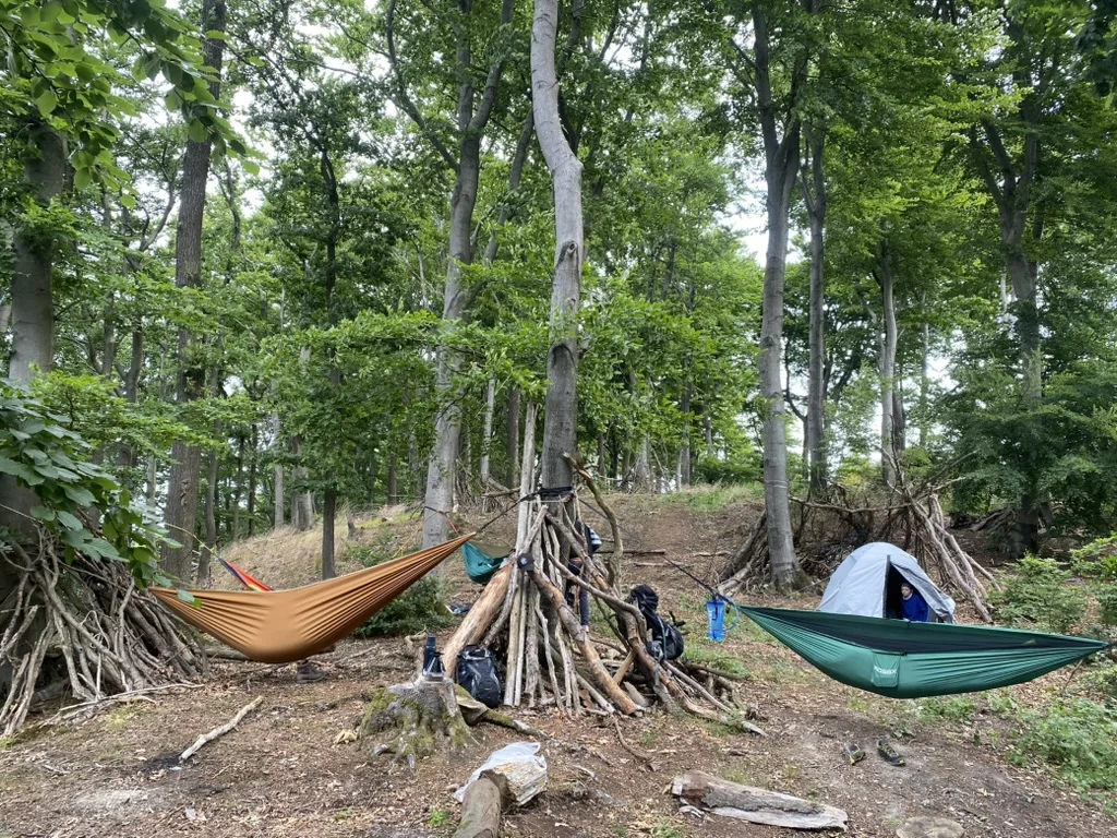 Zwei Hängematten und ein Zelt sind zwischen Bäumen im Wald aufgebaut. Rundherum stehen improvisierte Unterstände aus Ästen und mehrere Rucksäcke. | © DAV Dortmund
