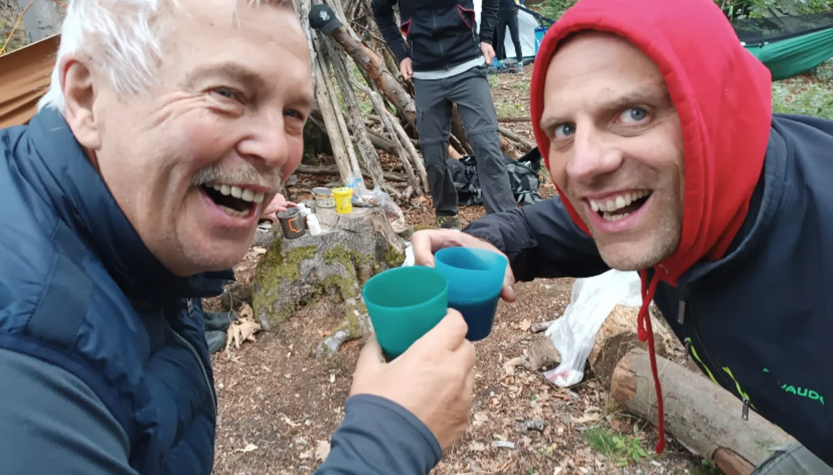 Zwei Männer stoßen mit bunten Campingbechern an und lachen fröhlich. Im Hintergrund sind Zelte, Rucksäcke und eine Gruppe von Menschen im Wald zu sehen. | © DAV Dortmund